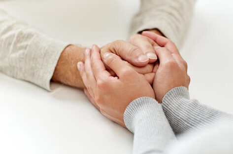 Die Hände einer älteren Person werden helfend von den Händen einer jüngeren Person umschlossen.