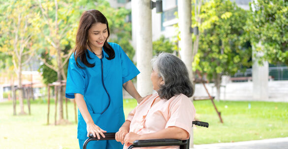 Eine Pflegerin steht mit einer Seniorin, die im Rollstuhl sitzt, im Garten eines Pflegeheimes und unterhält sich mit ihr.