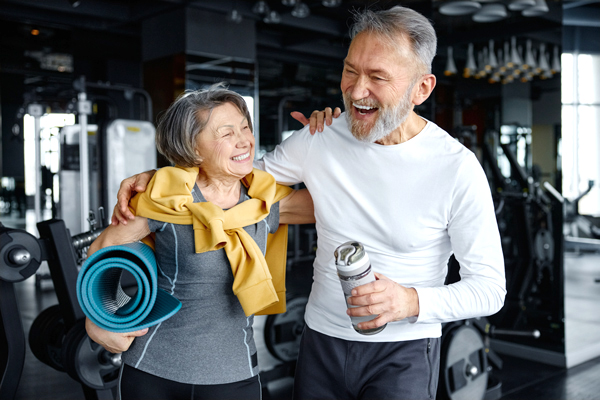 Ein Senior und eine Seniorin stehen im Fitnessstudio und lachen. Der Senior hat seine Trinkflasche in der Hand und hat den Arm um die Seniorin gelegt.