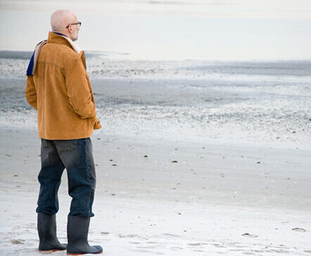 Ein Senior steht mit dem Rücken zum Betrachter und schaut auf das Meer hinaus.